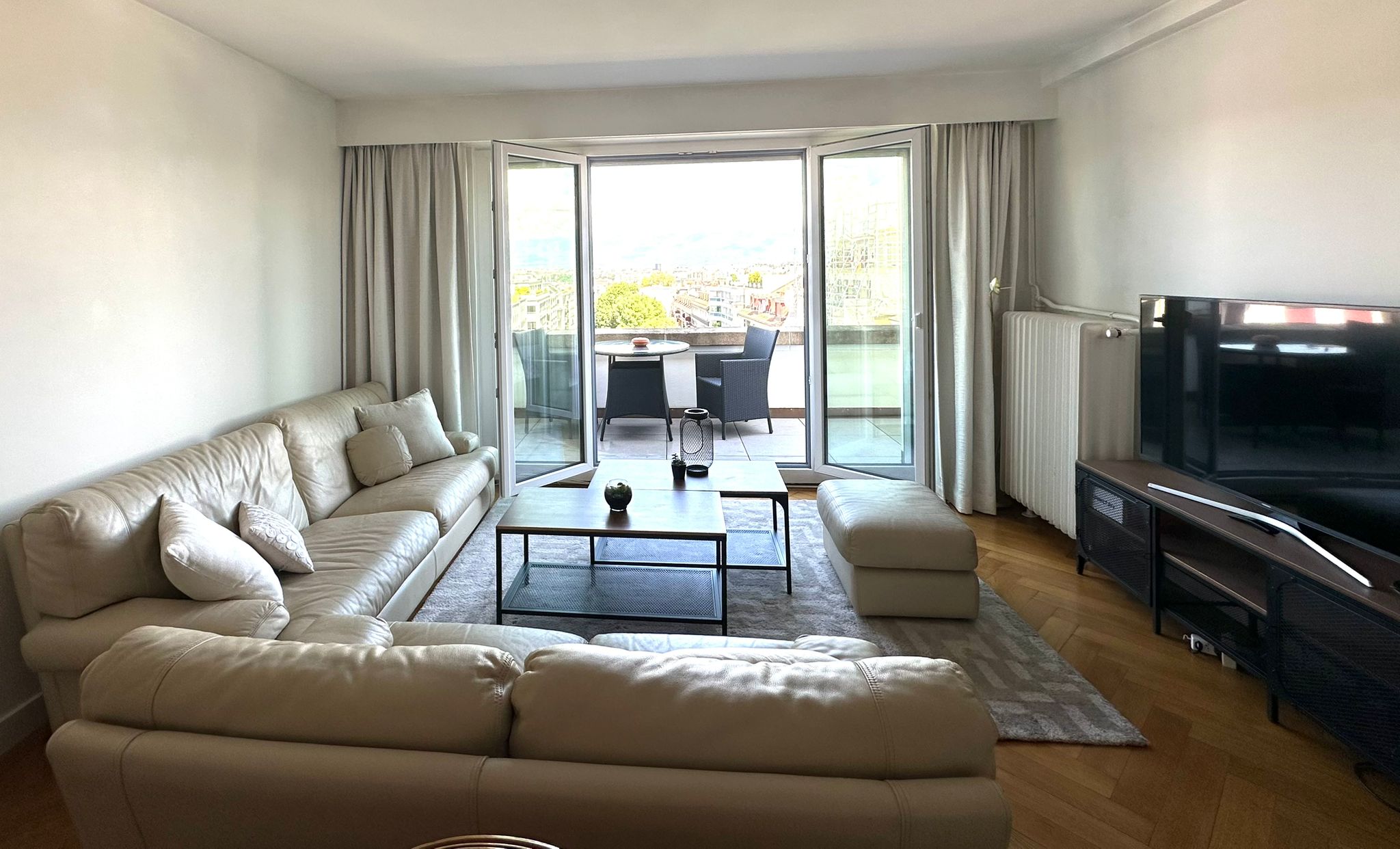 Spacieux salon avec une grande baie vitrée donnant sur le balcon et sa magnifique vue dégagée sur les montagnes et aperçu de jet d'eau, parquet en chevron